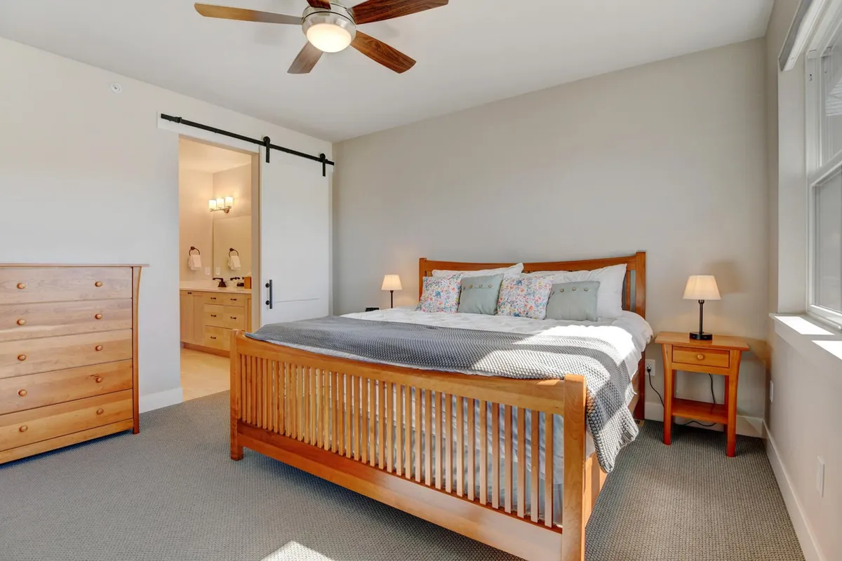 A serene bedroom featuring a wooden bed frame, colorful pillows, matching nightstands with warm lamps, and a dresser. Natural light filters in, creating a tranquil and cozy atmosphere ideal for relaxation or an afternoon nap.