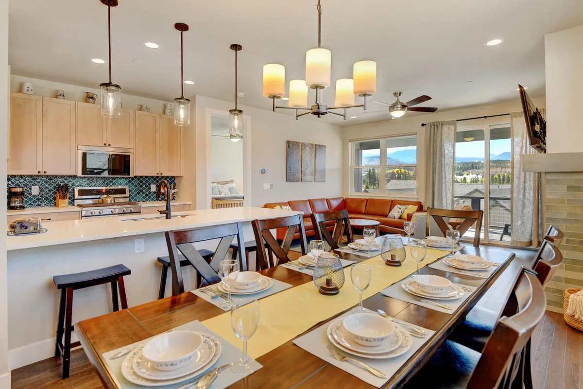 A view of the dining area with a custom wood table set for a meal, seamlessly adjacent to a modern kitchen featuring stainless steel appliances and granite countertops. Large windows reveal stunning mountain vistas, and the scene exudes a welcoming, communal atmosphere.