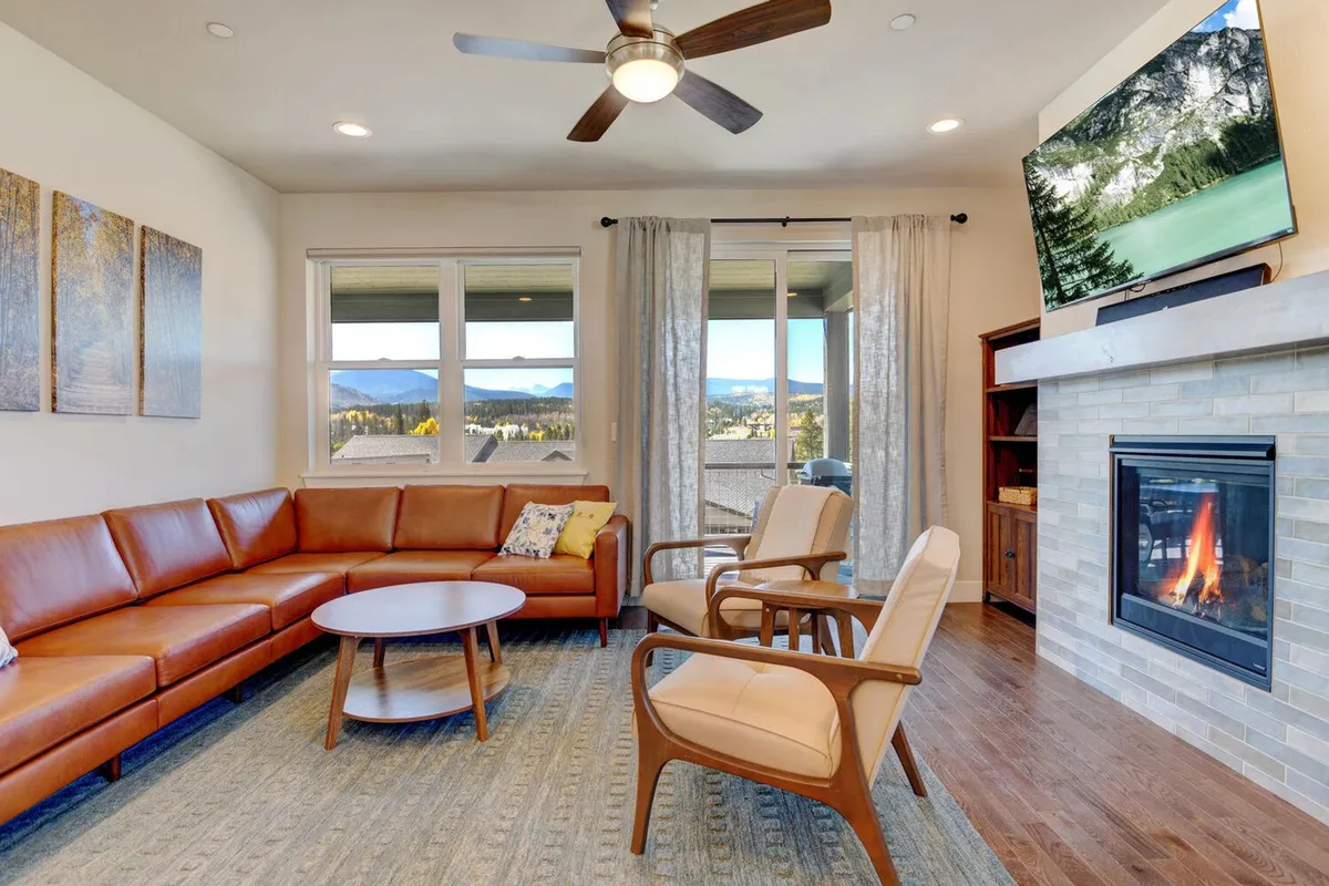 The image showcases a spacious, inviting living room featuring a luxurious leather sectional sofa arranged around a modern fireplace, with expansive windows framing mountain views. A round coffee table is set for gatherings, and a large TV completes the entertainment space, all bathed in natural light and designed for comfort and relaxation.