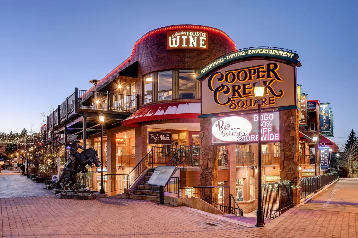 A lively exterior street scene featuring the Cooper Creek Square sign, boutique storefronts, outdoor seating, and decorative lighting, evoking the vibrant downtown Winter Park atmosphere.