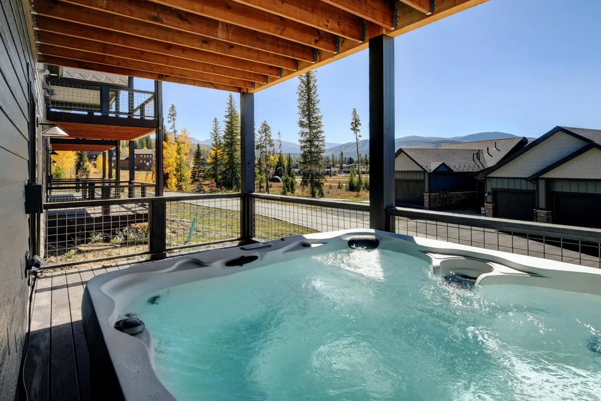 An outdoor hot tub sits on a private deck surrounded by tall trees, with sweeping mountain views in the background. The tub's bubbling water invites relaxation while the serene alpine landscape creates a sense of tranquility and escape.