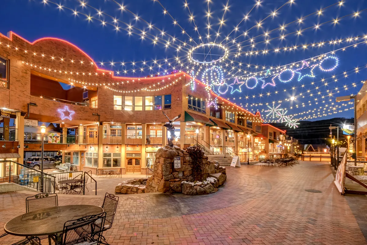 A vibrant exterior plaza scene featuring decorative string lights, a central fountain, and adjacent buildings. People gather to dine at outdoor cafes and shop at boutique stores, creating a lively, welcoming atmosphere.